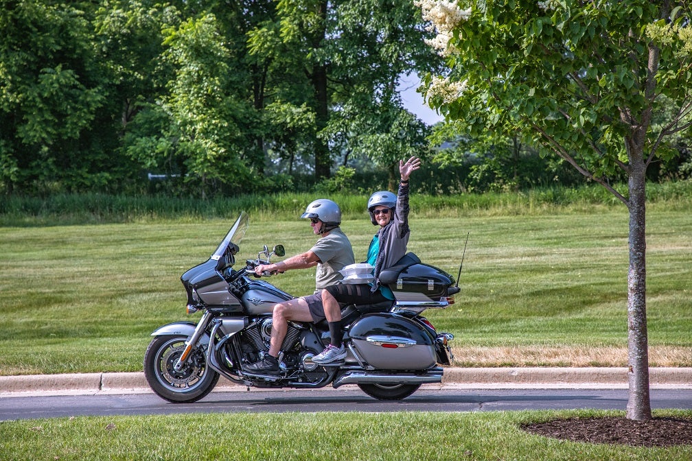 A couple rides away on a motorcycle as they depart the annual meeting. The passenger waves from the back.