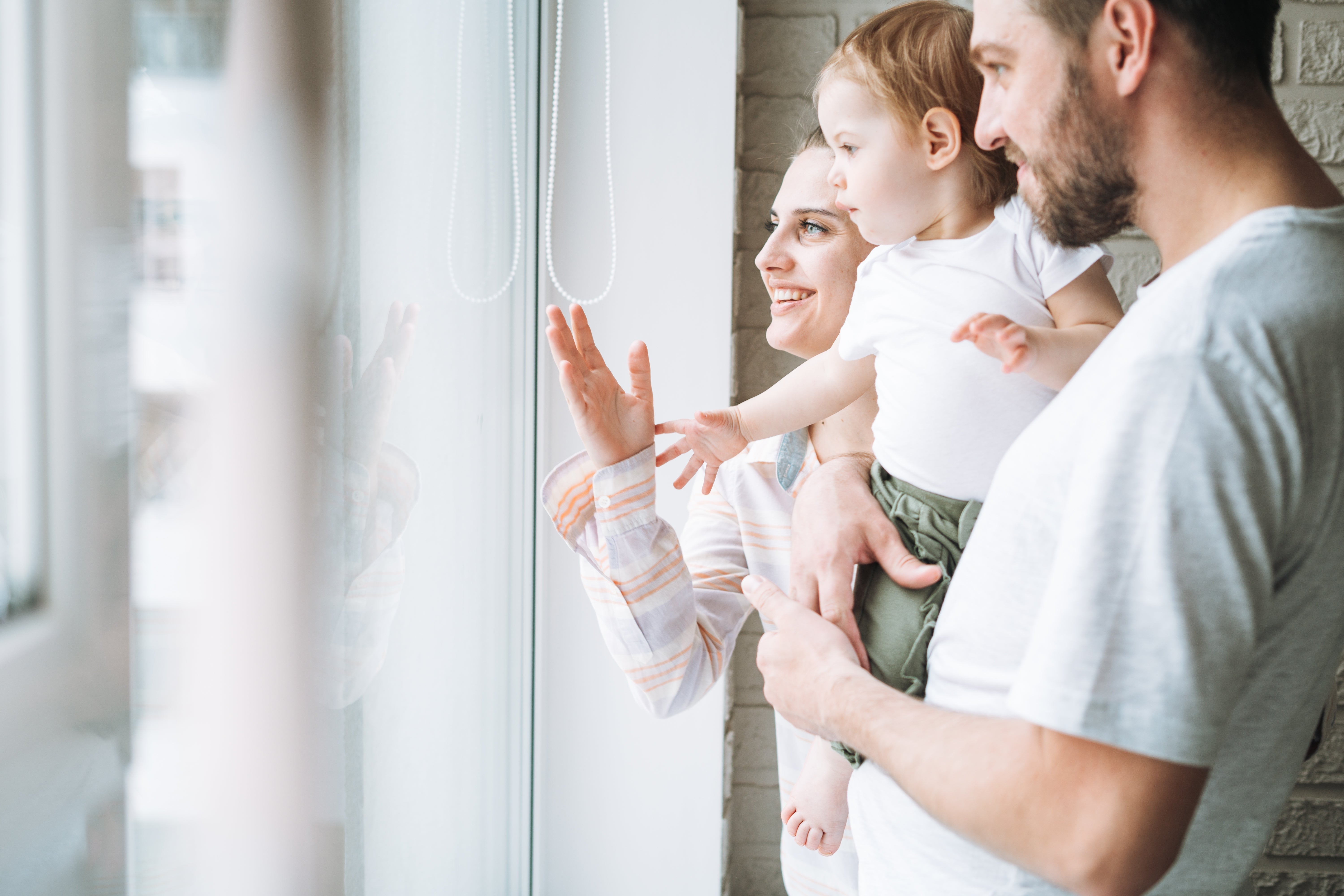A family stands smiling and looking out the window of their home.